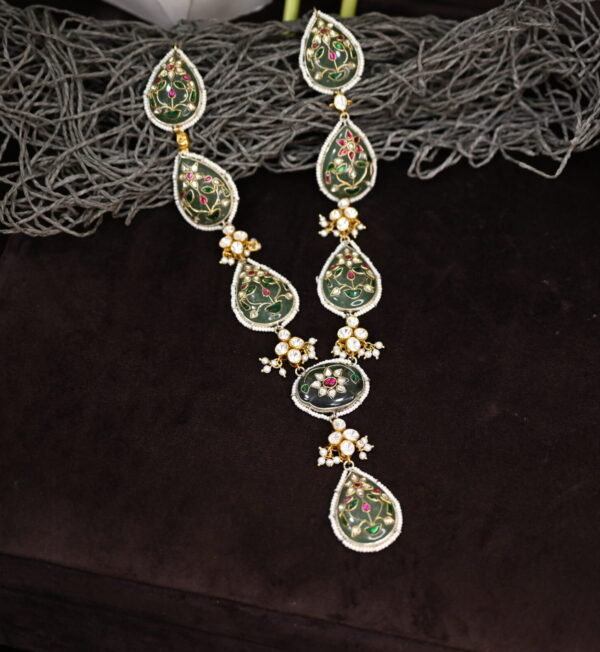 The image shows a necklace with a unique design. It has a central teardrop-shaped pendant with a flower motif, surrounded by smaller teardrop-shaped pendants. The necklace is made of gold and features intricate enamel work in a deep green color. It also has diamonds and pearls scattered throughout, adding to its sparkle and elegance.