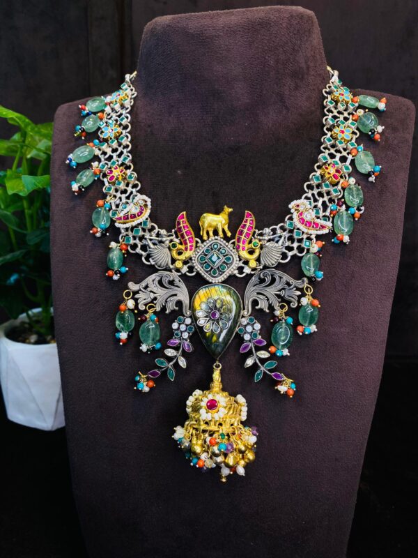 The image shows a beautiful necklace with a fusion of traditional and contemporary styles. Central Motif: The centerpiece features a large, ornate pendant with a blend of geometric patterns and floral motifs. It is adorned with a variety of colorful gemstones, including emeralds, rubies, and pearls.