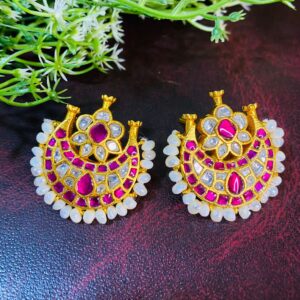 The image shows a pair of elegant Kundan earrings. The earrings are designed in a crescent moon shape with a floral motif at the center. The crescent is adorned with sparkling white Kundan stones and vibrant pink gemstones, while delicate pearls line the edges. The earrings are set against a backdrop of lush green foliage, enhancing their beauty.