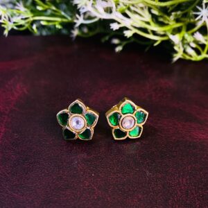 The image shows a pair of small, circular stud earrings. Each earring features a delicate floral design with five petals. The petals are embellished with small, green gemstones, and in the center of each flower is a single, round, white gemstone. The earrings appear to be made of gold or a gold-toned metal. They are resting on a dark, textured surface, possibly leather. A sprig of green foliage is also present in the image, adding a touch of nature.