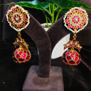 The image shows a pair of stunning earrings. They appear to be made of gold and feature intricate designs. The earrings have a circular base with a floral pattern and a dangling jhumka (drop) adorned with colorful stones. The jhumka has a tear-drop shape and is embellished with small pearls and colorful stones. The overall design gives a traditional Indian jewelry look.