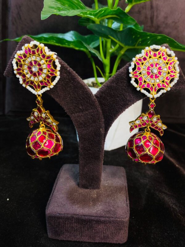 The image shows a pair of stunning earrings. They appear to be made of gold and feature intricate designs. The earrings have a circular base with a floral pattern and a dangling jhumka (drop) adorned with colorful stones. The jhumka has a tear-drop shape and is embellished with small pearls and colorful stones. The overall design gives a traditional Indian jewelry look.