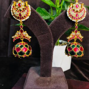 The image shows a pair of elegant Kundan earrings displayed on a jewelry stand. The earrings have a traditional Indian design with intricate detailing. The main part of the earring is a round shape with a red and green Meenakari design and is adorned with sparkling white Kundan stones. Below this, there is a smaller, teardrop-shaped pendant with a similar design and Kundan embellishments. The earrings are suspended from a delicate chain and hook. The background of the image is blurred and features a potted plant and a wooden surface.