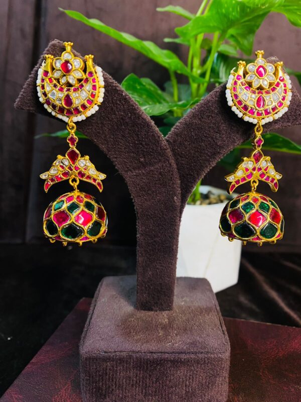The image shows a pair of elegant Kundan earrings displayed on a jewelry stand. The earrings have a traditional Indian design with intricate detailing. The main part of the earring is a round shape with a red and green Meenakari design and is adorned with sparkling white Kundan stones. Below this, there is a smaller, teardrop-shaped pendant with a similar design and Kundan embellishments. The earrings are suspended from a delicate chain and hook. The background of the image is blurred and features a potted plant and a wooden surface.