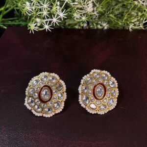 The image shows a pair of elegant Kundan earrings. The earrings feature a circular design with a floral motif, adorned with sparkling white Kundan stones. A touch of red Kundan adds a vibrant pop of color to the center, enhancing the overall elegance. The earrings are delicately crafted with gold-toned metal, adding to their luxurious appeal.
