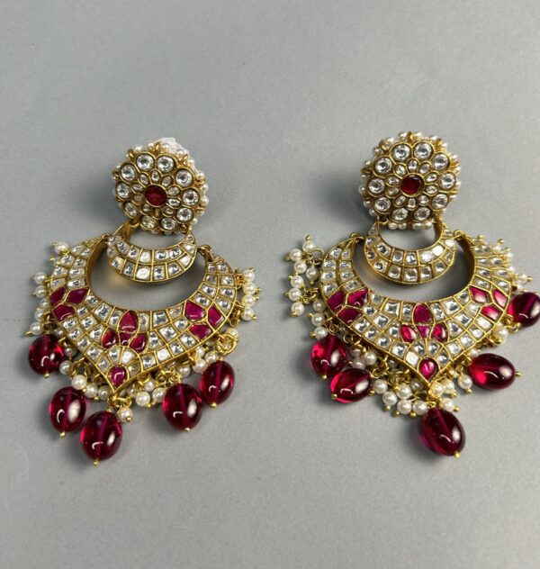 The image shows a pair of stunning red and white Kundan earrings. The earrings feature a circular base with a floral design, from which hangs a crescent-shaped pendant adorned with intricate Kundan work and white stones. The pendant is further embellished with delicate chains and red beads, creating a captivating cascade of colors. The overall design exudes a vintage charm and is sure to make a statement.