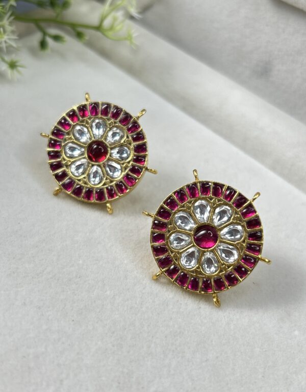 The image showcases a pair of exquisite Kundan earrings. The earrings are circular in shape and feature a central floral motif adorned with white Kundan stones. Surrounding the floral motif is a circular border of red Kundan stones, creating a striking contrast. The earrings are suspended from delicate gold-toned loops. The overall design evokes a sense of elegance and traditional Indian craftsmanship. "Kundan Spark Studs Earring"
