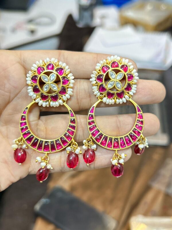 The image shows a pair of elegant Kundan earrings. The earrings feature a circular base adorned with white pearls, framing a central floral motif created with Kundan stones. A small diamond-like stone adds a touch of sparkle to the center. The earrings hang from a crescent-shaped element, also embellished with Kundan work and pearls. The crescent is further adorned with a row of pink beads, adding a pop of color. The earrings dangle with multiple strands of pink and white beads, completing the enchanting look.