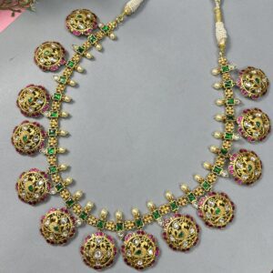 The image showcases a beautiful necklace adorned with intricate Kundan work. The necklace features a series of round pendants, each embellished with a peacock motif. The peacocks are crafted with delicate details, including their feathers and eyes. The necklace is further enhanced with sparkling green and pink gemstones, adding a touch of vibrant color. The overall design exudes a regal and sophisticated aesthetic. Gold