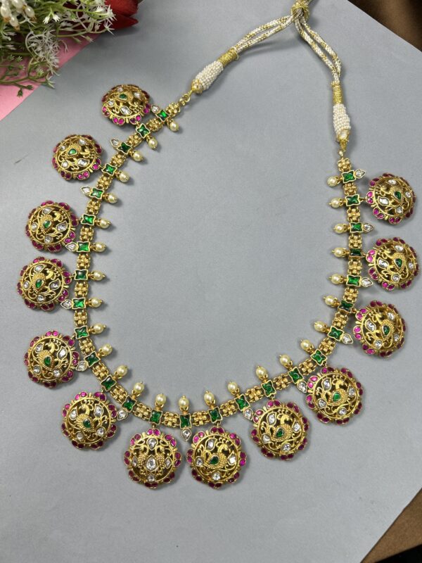 The image showcases a beautiful necklace adorned with intricate Kundan work. The necklace features a series of round pendants, each embellished with a peacock motif. The peacocks are crafted with delicate details, including their feathers and eyes. The necklace is further enhanced with sparkling green and pink gemstones, adding a touch of vibrant color. The overall design exudes a regal and sophisticated aesthetic. Gold