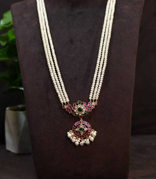 The image shows a beautiful necklace with a multi-strand pearl design. The necklace has a central pendant adorned with intricate Kundan work, featuring colorful gemstones like rubies and emeralds. The pendant also has delicate pearl embellishments. The necklace is displayed on a dark background with a hint of greenery in the corner.