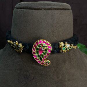 The image shows a close-up of a traditional Indian necklace. The necklace features a prominent paisley motif, a popular design element in Indian art and jewelry. The paisley is embellished with vibrant pink and green gemstones, likely set using the Kundan technique, where stones are encased in gold foil. The necklace is suspended on a black cord or thread.