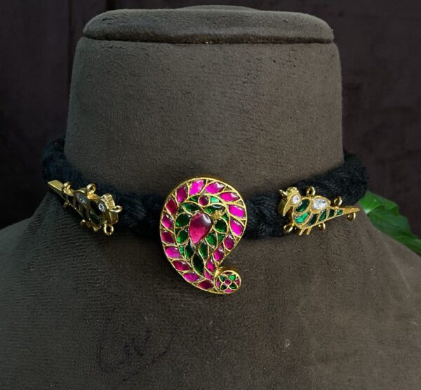 The image shows a close-up of a traditional Indian necklace. The necklace features a prominent paisley motif, a popular design element in Indian art and jewelry. The paisley is embellished with vibrant pink and green gemstones, likely set using the Kundan technique, where stones are encased in gold foil. The necklace is suspended on a black cord or thread.
