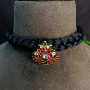 The image shows a close-up of a delicate necklace. It appears to be a black thread choker with a pendant featuring a lotus flower design. The lotus is embellished with vibrant pink and green kundan stones, creating a beautiful and eye-catching contrast against the black thread. The intricate craftsmanship and attention to detail are evident in the design.