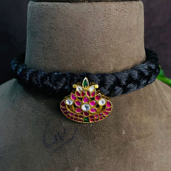 The image shows a close-up of a delicate necklace. It appears to be a black thread choker with a pendant featuring a lotus flower design. The lotus is embellished with vibrant pink and green kundan stones, creating a beautiful and eye-catching contrast against the black thread. The intricate craftsmanship and attention to detail are evident in the design.