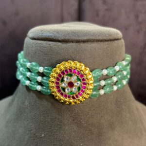 The image showcases a stunning choker necklace adorned with a vibrant green gemstone and a captivating floral Kundan pendant. The choker itself appears to be a multi-strand design, featuring alternating strands of green beads and pearls. The focal point of the necklace is the circular Kundan pendant, which is embellished with a mesmerizing array of colorful stones. The pendant features a central floral motif with delicate gold detailing, creating a truly exquisite piece of jewelry.