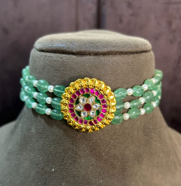 The image showcases a stunning choker necklace adorned with a vibrant green gemstone and a captivating floral Kundan pendant. The choker itself appears to be a multi-strand design, featuring alternating strands of green beads and pearls. The focal point of the necklace is the circular Kundan pendant, which is embellished with a mesmerizing array of colorful stones. The pendant features a central floral motif with delicate gold detailing, creating a truly exquisite piece of jewelry.
