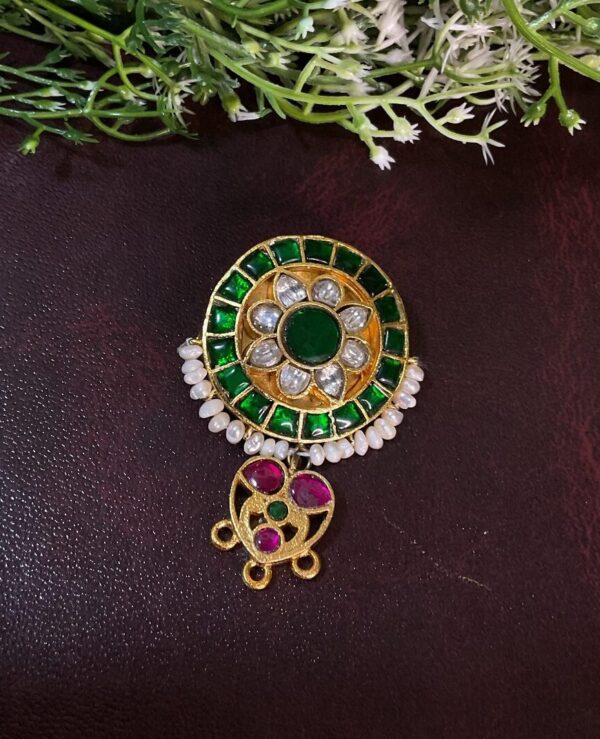 The image showcases a stunning Kundan pendant set against a dark background. The pendant features a circular design with a central, large Kundan stone surrounded by a halo of smaller green Kundan stones. The pendant is further embellished with delicate pearls and ruby accents, adding to its overall charm.