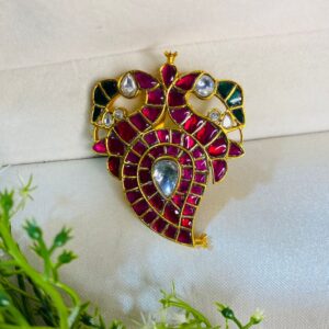 The pendant seems to be resting on a bed of green foliage, possibly a plant or a piece of cloth. The lighting in the image is well-balanced, enhancing the colors and details of the pendant. Intricate Ruby Kundan Pendant