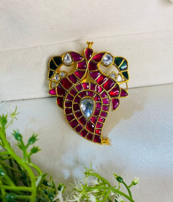 The pendant seems to be resting on a bed of green foliage, possibly a plant or a piece of cloth. The lighting in the image is well-balanced, enhancing the colors and details of the pendant. Intricate Ruby Kundan Pendant
