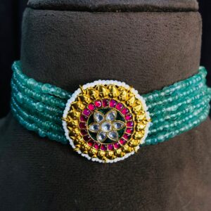 The image shows a beautiful choker necklace made with green beads. The choker has multiple strands of beads and features a large, circular pendant in the center. The pendant is adorned with Kundan work, which involves setting precious and semi-precious stones in gold foil. The pendant has a floral design with a central stone surrounded by smaller colorful stones. Multi-Strand