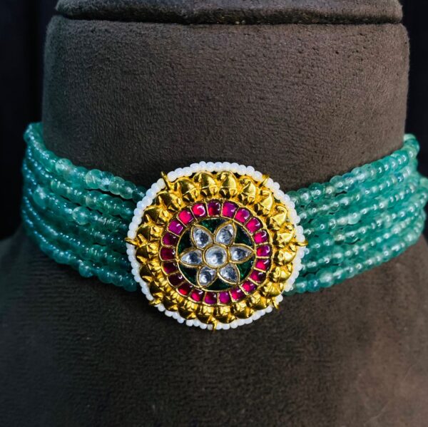The image shows a beautiful choker necklace made with green beads. The choker has multiple strands of beads and features a large, circular pendant in the center. The pendant is adorned with Kundan work, which involves setting precious and semi-precious stones in gold foil. The pendant has a floral design with a central stone surrounded by smaller colorful stones. Multi-Strand