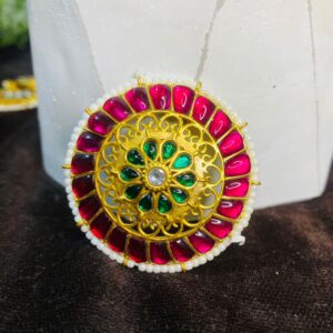 The image shows a close-up of a traditional Indian headpiece called a Maangtika. It is a circular ornament, likely made of gold or a gold-toned metal, and is adorned with intricate floral patterns. The Maangtika is embellished with colorful gemstones, possibly rubies, emeralds, and diamonds, creating a dazzling display of colors. The overall design suggests a rich cultural heritage and showcases the exquisite craftsmanship of Indian jewelry-making.