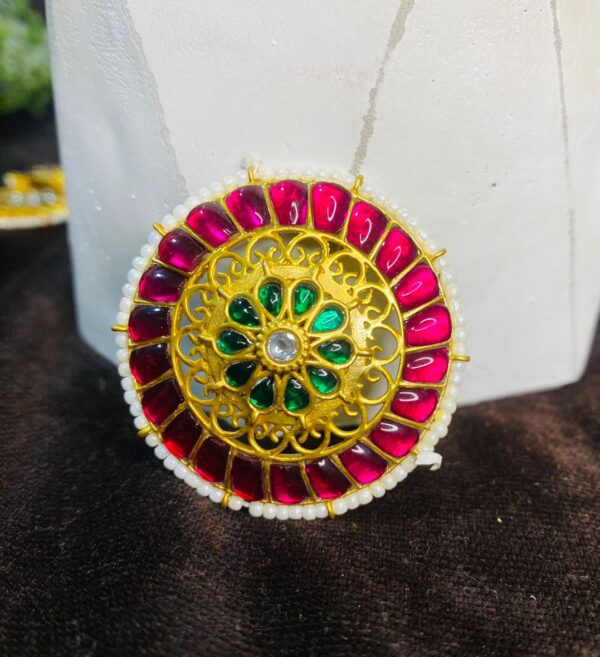 The image shows a close-up of a traditional Indian headpiece called a Maangtika. It is a circular ornament, likely made of gold or a gold-toned metal, and is adorned with intricate floral patterns. The Maangtika is embellished with colorful gemstones, possibly rubies, emeralds, and diamonds, creating a dazzling display of colors. The overall design suggests a rich cultural heritage and showcases the exquisite craftsmanship of Indian jewelry-making.