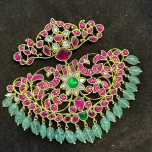 The image showcases a stunning Kundan pendant. The pendant features a large, crescent-shaped base adorned with intricate floral motifs and embellished with a profusion of colorful gemstones. The dominant colors are ruby and emerald, with diamonds adding sparkle and enhancing the overall brilliance. The pendant exudes a regal charm and would make a perfect statement piece for special occasions. The Kundan work, a traditional Indian technique of setting gemstones in gold foil, creates a mesmerizing three-dimensional effect.