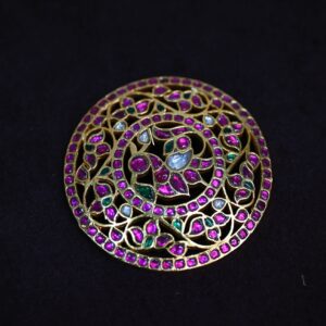 The image appears to be a close-up shot of a circular pendant or brooch. It showcases intricate craftsmanship with a prominent circular motif adorned with numerous small, colorful gemstones. The gemstones are set in a delicate pattern, possibly creating a floral or geometric design. The overall color scheme is a mix of vibrant hues, likely including pink, green, and perhaps white or gold.