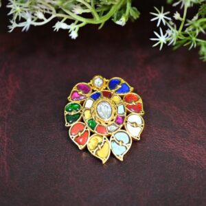 The image shows a beautiful Kundan pendant. It has a circular design with a central white stone, possibly a diamond or a high-quality imitation, surrounded by a colorful array of stones. The colors include vibrant reds, blues, greens, and yellows, creating a mesmerizing mosaic-like pattern. The pendant is set in a delicate gold frame, showcasing intricate craftsmanship and meticulous attention to detail. The overall impression is one of luxury and regal charm.
