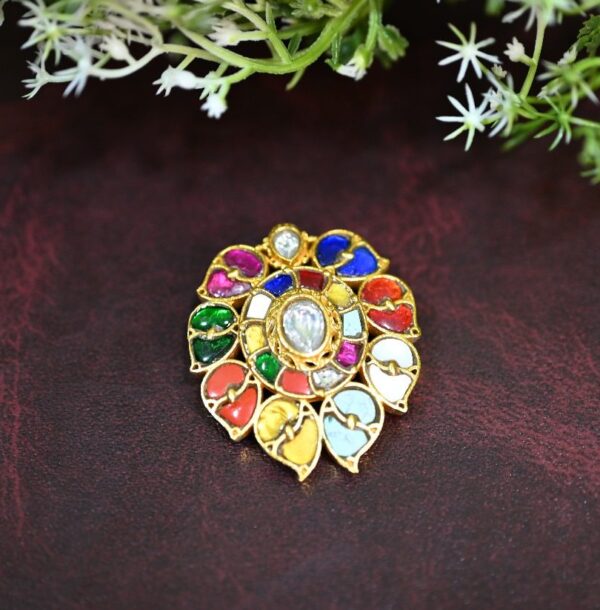 The image shows a beautiful Kundan pendant. It has a circular design with a central white stone, possibly a diamond or a high-quality imitation, surrounded by a colorful array of stones. The colors include vibrant reds, blues, greens, and yellows, creating a mesmerizing mosaic-like pattern. The pendant is set in a delicate gold frame, showcasing intricate craftsmanship and meticulous attention to detail. The overall impression is one of luxury and regal charm.