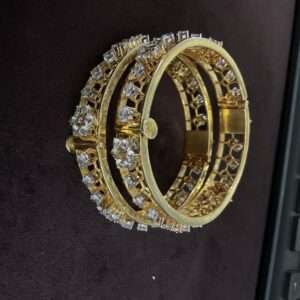 The image shows a pair of moissanite bangles. The bangles are circular and appear to be made of silver. They are adorned with numerous small, clear stones, presumably moissanites, which sparkle and catch the light. The bangles are stacked on top of each other, with the lower bangle slightly larger than the upper one. The background is a dark, textured surface, possibly a fabric or leather.