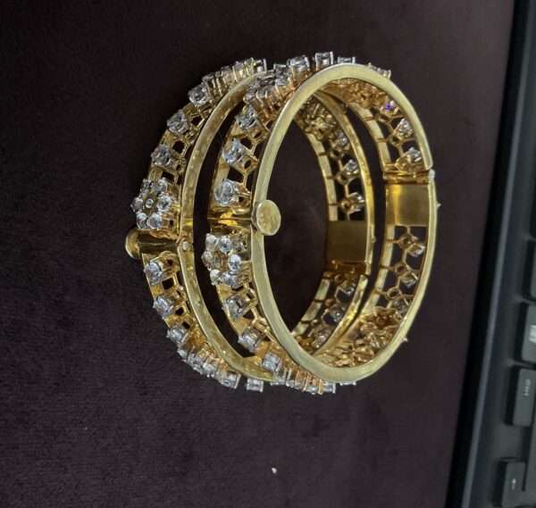 The image shows a pair of moissanite bangles. The bangles are circular and appear to be made of silver. They are adorned with numerous small, clear stones, presumably moissanites, which sparkle and catch the light. The bangles are stacked on top of each other, with the lower bangle slightly larger than the upper one. The background is a dark, textured surface, possibly a fabric or leather.
