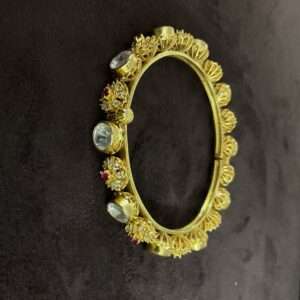 The image shows a pair of gold-plated bangles with intricate designs and embedded gemstones. The bangles appear to be made of metal, possibly brass or copper, and are plated with a layer of gold. The designs on the bangles are elaborate, featuring floral motifs, geometric patterns, and small, colorful gemstones. The gemstones are likely cubic zirconia or other synthetic stones, adding sparkle and color to the overall design. The bangles are wide and have a slightly curved shape, suggesting they are meant to be worn on the upper arm. The background is dark, making the gold plating and gemstones stand out.