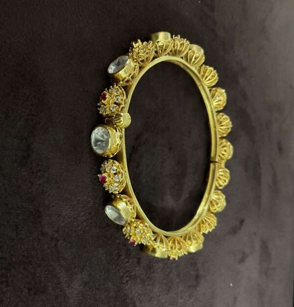 The image shows a pair of gold-plated bangles with intricate designs and embedded gemstones. The bangles appear to be made of metal, possibly brass or copper, and are plated with a layer of gold. The designs on the bangles are elaborate, featuring floral motifs, geometric patterns, and small, colorful gemstones. The gemstones are likely cubic zirconia or other synthetic stones, adding sparkle and color to the overall design. The bangles are wide and have a slightly curved shape, suggesting they are meant to be worn on the upper arm. The background is dark, making the gold plating and gemstones stand out.