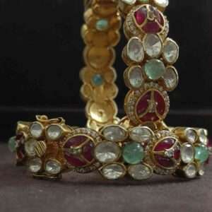 The image shows a pair of elegant and ornate bangles. They appear to be made of gold or a similar metallic material, encrusted with a variety of colorful gemstones. There are large, round stones in shades of green and red, as well as smaller, white stones that might be diamonds or another type of colorless gemstone.
