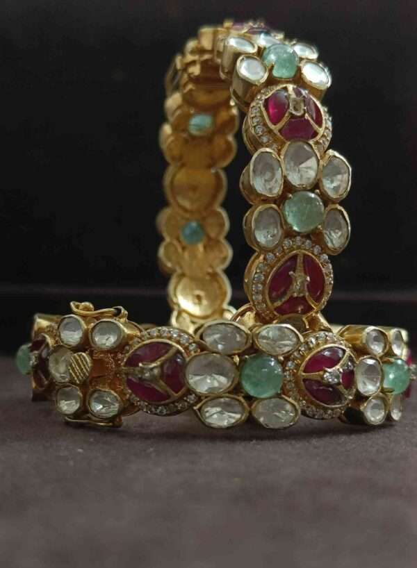 The image shows a pair of elegant and ornate bangles. They appear to be made of gold or a similar metallic material, encrusted with a variety of colorful gemstones. There are large, round stones in shades of green and red, as well as smaller, white stones that might be diamonds or another type of colorless gemstone.