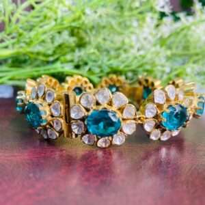 The image shows a pair of gold-toned bangles adorned with sparkling gemstones. The bangles appear to be made of metal, possibly silver or gold-plated, and feature a floral design with alternating oval-shaped green stones and clusters of smaller white stones. The green stones are likely Moissanite, known for their brilliance and fire.