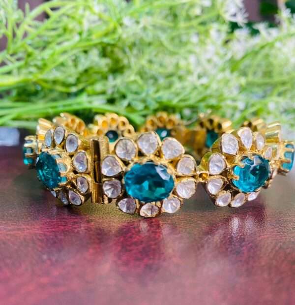 The image shows a pair of gold-toned bangles adorned with sparkling gemstones. The bangles appear to be made of metal, possibly silver or gold-plated, and feature a floral design with alternating oval-shaped green stones and clusters of smaller white stones. The green stones are likely Moissanite, known for their brilliance and fire.