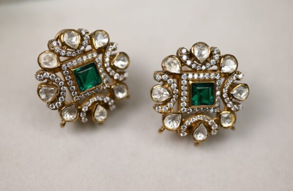 The image shows a pair of stunning earrings. They are made of gold and feature a square-shaped emerald gemstone in the center. The emerald is surrounded by a delicate floral design composed of small white stones, likely diamonds or moissanites. The earrings have a vintage or antique look and are sure to make a statement.