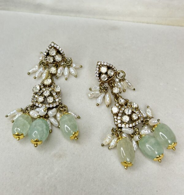 The image shows a pair of elegant and luxurious earrings. They have a multi-tiered design with each tier showcasing sparkling moissanites. The earrings are further embellished with delicate pearls and a cluster of beautiful green stones, adding a touch of color and contrast. The intricate craftsmanship and attention to detail make these earrings a true masterpiece. They would be perfect for special occasions or to add a touch of glamour to any outfit.