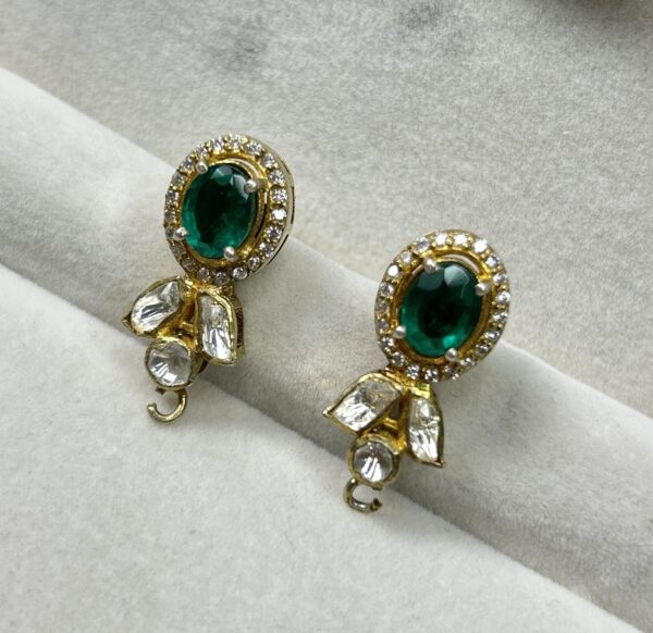 Center Stone: Each earring has a large, oval-shaped green gemstone, likely an emerald. Setting: The emerald is set in a gold-colored metal with a halo of smaller, round white gemstones. This halo adds a touch of sparkle and dimension. Design: The earrings have a delicate, feminine design with intricate details. They might have a dangling element or a small motif below the main stone.