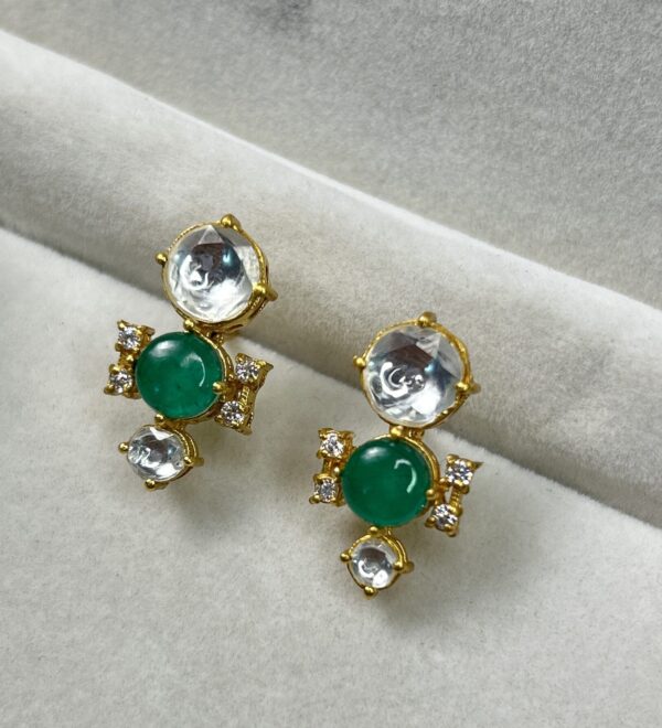 Center Stone: Each earring has a large, oval-shaped green gemstone, likely an emerald. Setting: The emerald is set in a gold-colored metal with a halo of smaller, round white gemstones. This halo adds a touch of sparkle and dimension. Design: The earrings have a delicate, feminine design with intricate details. They might have a dangling element or a small motif below the main stone.