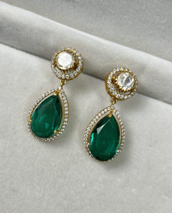The image shows a pair of elegant earrings on a white surface. The earrings are made of gold and feature a combination of green and white gemstones. Each earring has a teardrop-shaped green stone, surrounded by a halo of smaller white stones. Above the green stone, there is a circular cluster of white stones.