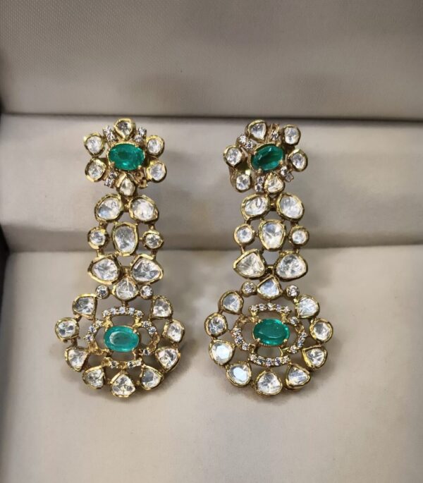 The image shows a pair of stunning dangle earrings. The earrings are made of gold and feature a cluster of round-cut diamonds and emeralds. The emeralds are a vibrant green and the diamonds sparkle brightly.