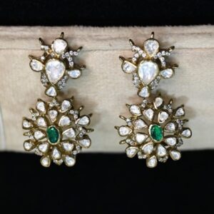 The image shows a pair of stunning, large drop earrings. They appear to be made of gold with a generous amount of sparkling white gemstones, likely diamonds or moissanites, and several vibrant green gemstones, likely emeralds or similar. The earrings feature intricate designs with multiple layers and a cascading effect. They are displayed on a black jewelry stand.