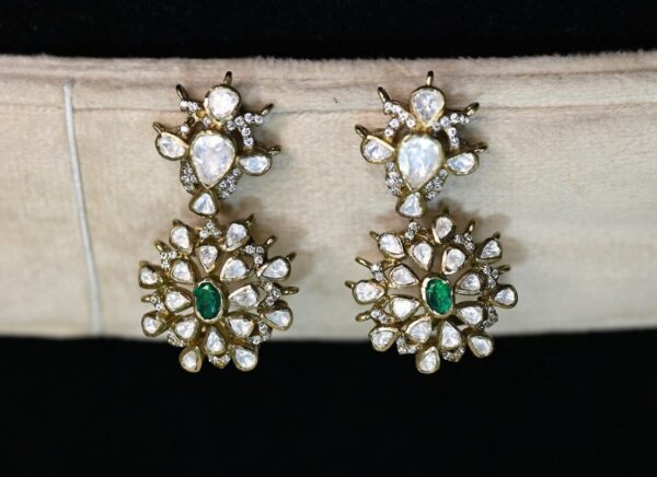 The image shows a pair of stunning, large drop earrings. They appear to be made of gold with a generous amount of sparkling white gemstones, likely diamonds or moissanites, and several vibrant green gemstones, likely emeralds or similar. The earrings feature intricate designs with multiple layers and a cascading effect. They are displayed on a black jewelry stand.
