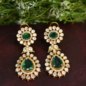 The image shows a pair of ornate earrings featuring a floral design. Each earring has two tiers: the top tier is a circular arrangement of small, clear gemstones around a central, deep green gemstone. The bottom tier is a similar arrangement but larger, with a pear-shaped green gemstone at the center. The earrings are gold in color and rest on a dark, textured surface. A sprig of small white flowers is positioned in the background, adding a touch of nature to the composition. Sources and related content