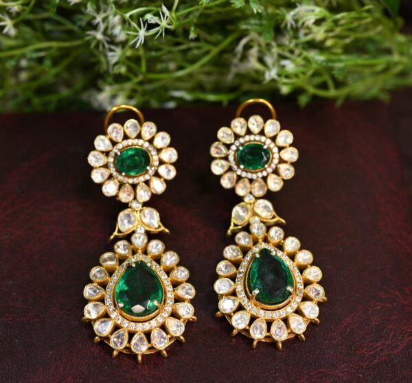 The image shows a pair of ornate earrings featuring a floral design. Each earring has two tiers: the top tier is a circular arrangement of small, clear gemstones around a central, deep green gemstone. The bottom tier is a similar arrangement but larger, with a pear-shaped green gemstone at the center. The earrings are gold in color and rest on a dark, textured surface. A sprig of small white flowers is positioned in the background, adding a touch of nature to the composition. Sources and related content